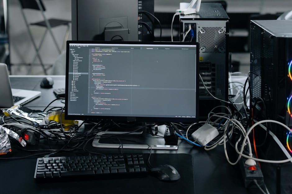 The image displays a cluttered workspace with a computer monitor at the center showing lines of code, surrounded by tangled cables and electronic devices, including a desktop tower with visible RGB li