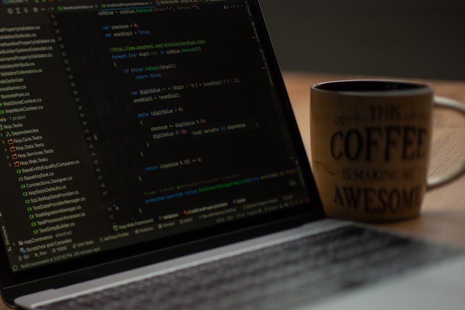 A close-up view of a laptop displaying lines of colorful programming code on its screen, placed on a wooden desk. Next to the laptop is a ceramic coffee mug with the phrase 'This coffee is making me a